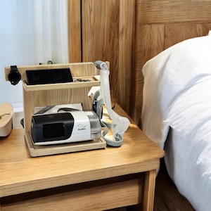 May include: A wooden bedside table with a white and gray CPAP machine and a hose on a shelf. The table is next to a bed with a white pillow and a white blanket.