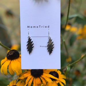 May include: A pair of black lightning bolt earrings hanging from a white card with the text "MamaTried" and "Nooga". The earrings are displayed against a backdrop of yellow and black flowers.