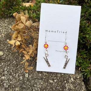 May include: A pair of handmade earrings featuring a red and yellow flower design above a silver clothespin charm. The earrings hang from silver hooks. The card behind the earrings reads "MamaTried" and "NoogaDood".
