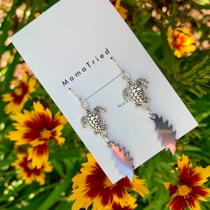 May include: Silver-toned earrings featuring a turtle charm and a jagged, lightning bolt-shaped pendant. The earrings hang from a simple hook. The background includes yellow and red flowers and a white card with the text "MamaTried".