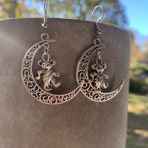 May include: Silver crescent moon earrings with a dancing bear charm hanging from each moon. The earrings have a filigree design.