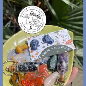 May include: A collection of witchy wares including a small bottle filled with black stones, a bottle filled with amethyst crystals, a bottle filled with green stones, a small mushroom charm, a small red mushroom charm, a small pink mushroom charm, a small orange mushroom charm, a small yellow mushroom charm, a small orange and red charm, a small blue charm, a small blue charm, and a small card that says "Custom Witchy Wares".