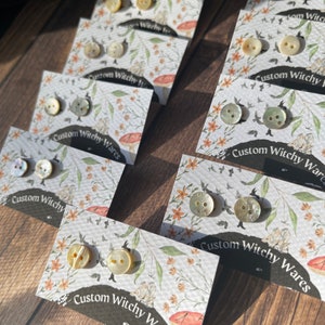 May include: Assortment of small, round, iridescent buttons displayed on cards with floral designs. The cards have the text "Custom Witchy Wares" printed on them.