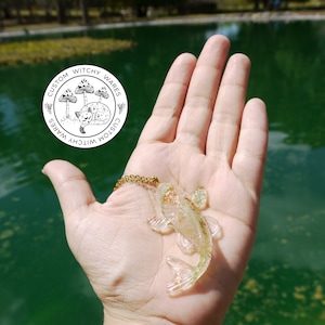 May include: A gold chain necklace with a clear resin pendant shaped like a fish. The fish has a glittery, iridescent finish and is set against a green background.