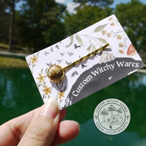 May include: A gold hair clip with a brown and white stone accent. The clip is on a white card with the text "Custom Witchy Wares".