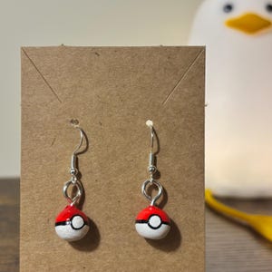 Puede incluir: Un par de pendientes hechos a mano con un diseño rojo y blanco con una franja negra, que se asemeja a una Poké Ball. Los pendientes tienen ganchos plateados y se exhiben sobre un soporte de cartón marrón.