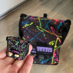 May include: A small black bag and a larger black bag with a colourful splatter paint design. The small bag has the word "ORGANIC" printed on it. The bags have a black leather trim.