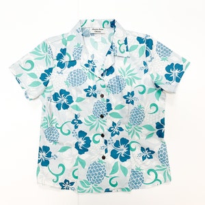 May include: A white short-sleeved button-down shirt with a blue and green pineapple and floral print. The shirt has a collar and a button closure.