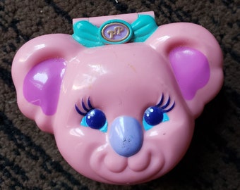 May include: Pink plastic koala-shaped compact with a turquoise bow and a gold button with the letters "PT" on it. The koala has purple ears, blue eyes with eyelashes, and a purple nose.