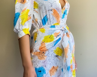 Puede incluir: Un vestido blanco mini con estampado floral abstracto azul, amarillo y naranja. El vestido tiene un escote en V, mangas 3/4 y un lazo en la cintura.