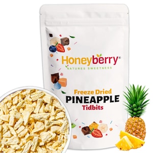 May include: A white stand-up pouch labelled Honeyberry Freeze Dried PINEAPPLE Tidbits, featuring illustrations of fruit and chocolate. Beside the pouch is a bowl filled with light yellow, freeze-dried pineapple pieces and a whole pineapple.