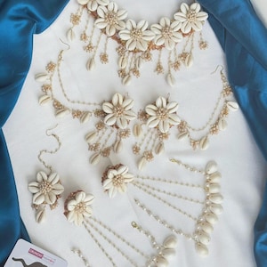 May include: A jewellery set featuring white cowrie shells and small beads arranged into flower shapes. The set includes a necklace, a pair of earrings, a headpiece, and a hand ornament, all connected with thin gold-toned chains and dangling shell accents.