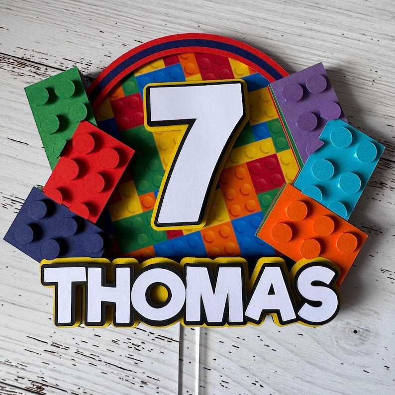 Op de afbeelding: Een kleurrijke Lego-thema caketopper met een wit nummer 7 en de naam "THOMAS" in witte letters. De topper is versierd met kleurrijke Lego-stenen.