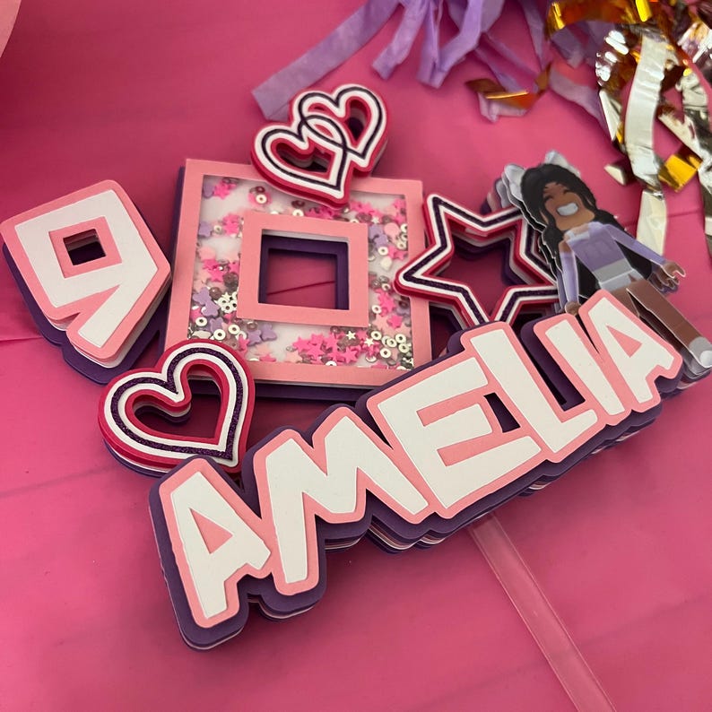 May include: A pink and purple cake topper with the name Amelia, a number 9, a heart, a star, and a Roblox character. The topper is decorated with glitter.