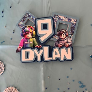 May include: A blue and white cake topper with the name "DYLAN" and the number "9" in the center. Two Roblox characters are on either side of the number. The characters are wearing blue and red shirts with the word "ROBLOX" on them.