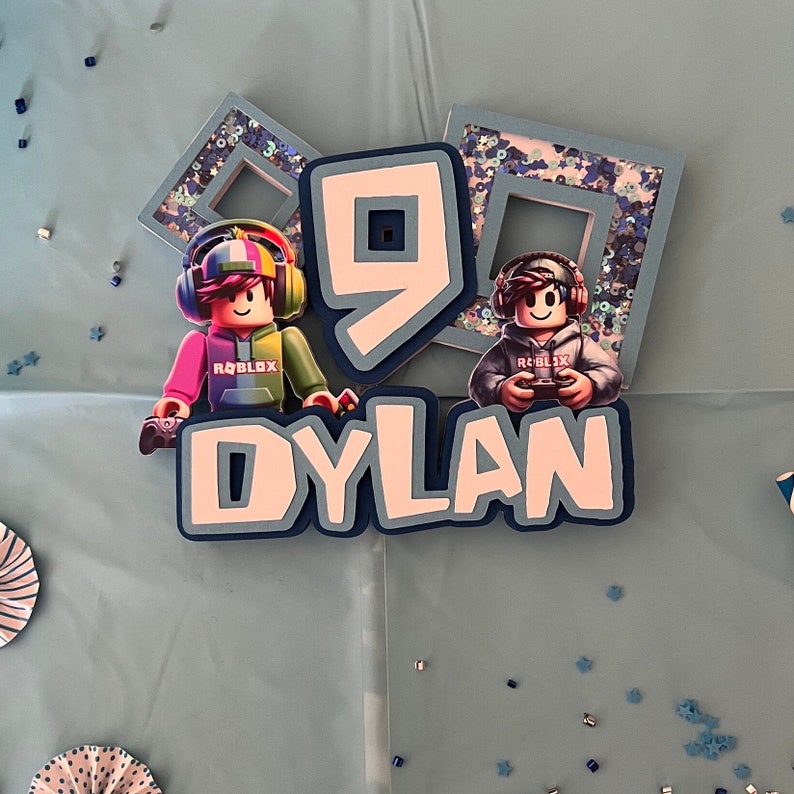 May include: A blue and white cake topper with the name "DYLAN" and the number "9" in the center. Two Roblox characters are on either side of the number. The characters are wearing blue and red shirts with the word "ROBLOX" on them.