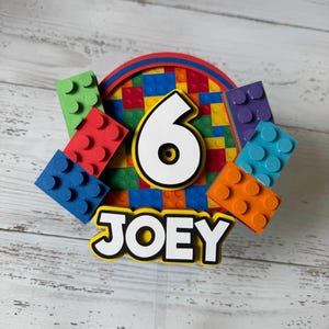 Op de afbeelding: Een kleurrijke taarttopper met het nummer 6, de naam "JOEY" en een Lego-thema. De topper heeft een cirkel van kleurrijke Lego-stenen, met een wit nummer 6 in het midden. Er zijn ook verschillende individuele Lego-stenen rond de cirkel, in verschillende kleuren.