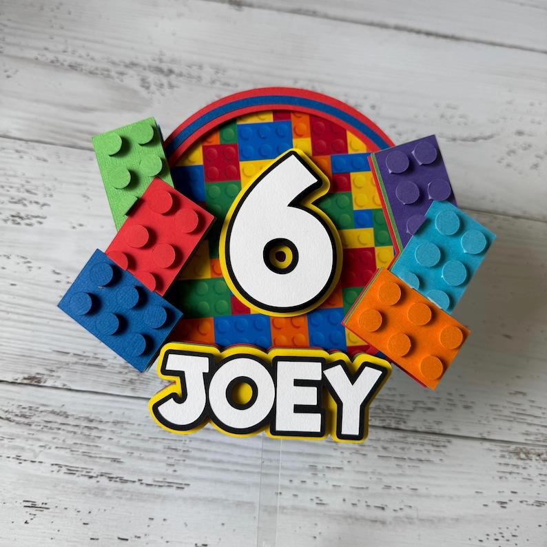 Op de afbeelding: Een kleurrijke taarttopper met het nummer 6, de naam "JOEY" en een Lego-thema. De topper heeft een cirkel van kleurrijke Lego-stenen, met een wit nummer 6 in het midden. Er zijn ook verschillende individuele Lego-stenen rond de cirkel, in verschillende kleuren.