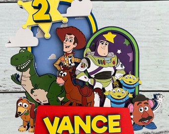 Pode incluir: Um topo de bolo em camadas com personagens de Toy Story, incluindo Woody, Buzz Lightyear, Rex, Bullseye, Slinky Dog, Sr. Cabeça de Batata e dois alienígenas. O design inclui uma estrela amarela com o número 2 e uma base vermelha com o nome VANCE.