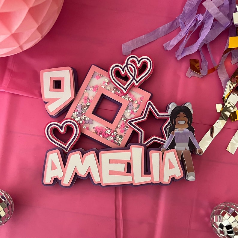 May include: Pink and white birthday decorations with the name Amelia, a pink and white number 9, a pink and white heart, a pink and white star, and a pink and white square with glitter. The decorations are on a pink background.