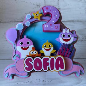 Peut inclure: D&eacute;coration de g&acirc;teau multicouche avec des requins de dessins anim&eacute;s, une &eacute;toile de mer et des bulles. Le chiffre "2" est en haut, avec le nom "SOFIA" en dessous. Le design incorpore des couleurs rose, violet, jaune et bleu, parfait pour un anniversaire.