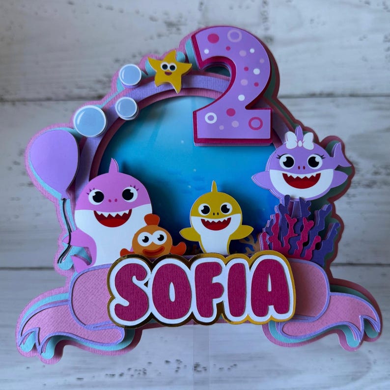 Peut inclure: D&eacute;coration de g&acirc;teau multicouche avec des requins de dessins anim&eacute;s, une &eacute;toile de mer et des bulles. Le chiffre "2" est en haut, avec le nom "SOFIA" en dessous. Le design incorpore des couleurs rose, violet, jaune et bleu, parfait pour un anniversaire.