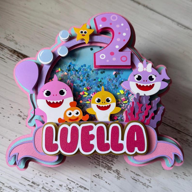 Peut inclure: D&eacute;coration de g&acirc;teau en papier superpos&eacute; avec des personnages de requins de dessins anim&eacute;s, une &eacute;toile de mer et des bulles. Le chiffre 2 est en haut et le nom "LUELLA" en bas. La palette de couleurs comprend le rose, le violet, le jaune et le bleu.