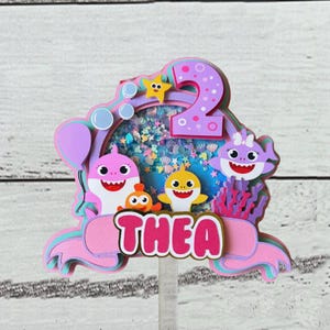 Peut inclure: D&eacute;coration de g&acirc;teau &agrave; plusieurs niveaux avec des requins de dessins anim&eacute;s, une &eacute;toile et le chiffre 2. Le nom "THEA" est affich&eacute; en lettres roses. La d&eacute;coration a une palette de couleurs rose, violet et bleu, avec un centre transparent rempli de confettis.
