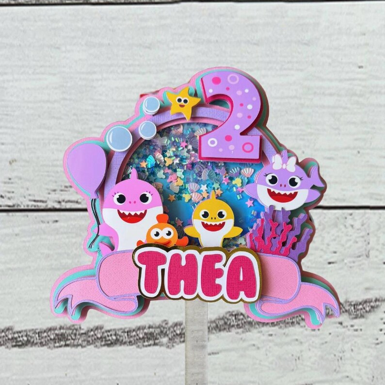 Peut inclure: D&eacute;coration de g&acirc;teau &agrave; plusieurs niveaux avec des requins de dessins anim&eacute;s, une &eacute;toile et le chiffre 2. Le nom "THEA" est affich&eacute; en lettres roses. La d&eacute;coration a une palette de couleurs rose, violet et bleu, avec un centre transparent rempli de confettis.