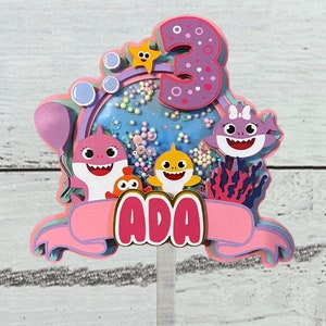 Peut inclure: D&eacute;coration de g&acirc;teau multicouche avec des personnages de requins de dessins anim&eacute;s, une &eacute;toile de mer et le chiffre 3. La d&eacute;coration est rose et violette avec le nom "ADA" sur une banni&egrave;re. Des bulles et des confettis sont &eacute;galement inclus.