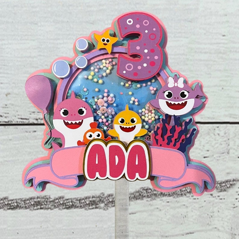 Peut inclure: D&eacute;coration de g&acirc;teau multicouche avec des personnages de requins de dessins anim&eacute;s, une &eacute;toile de mer et le chiffre 3. La d&eacute;coration est rose et violette avec le nom "ADA" sur une banni&egrave;re. Des bulles et des confettis sont &eacute;galement inclus.