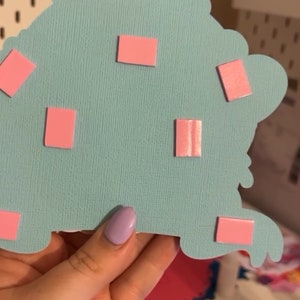 May include: A light blue paper craft with pink adhesive squares attached to the back. The craft is being held by a hand.