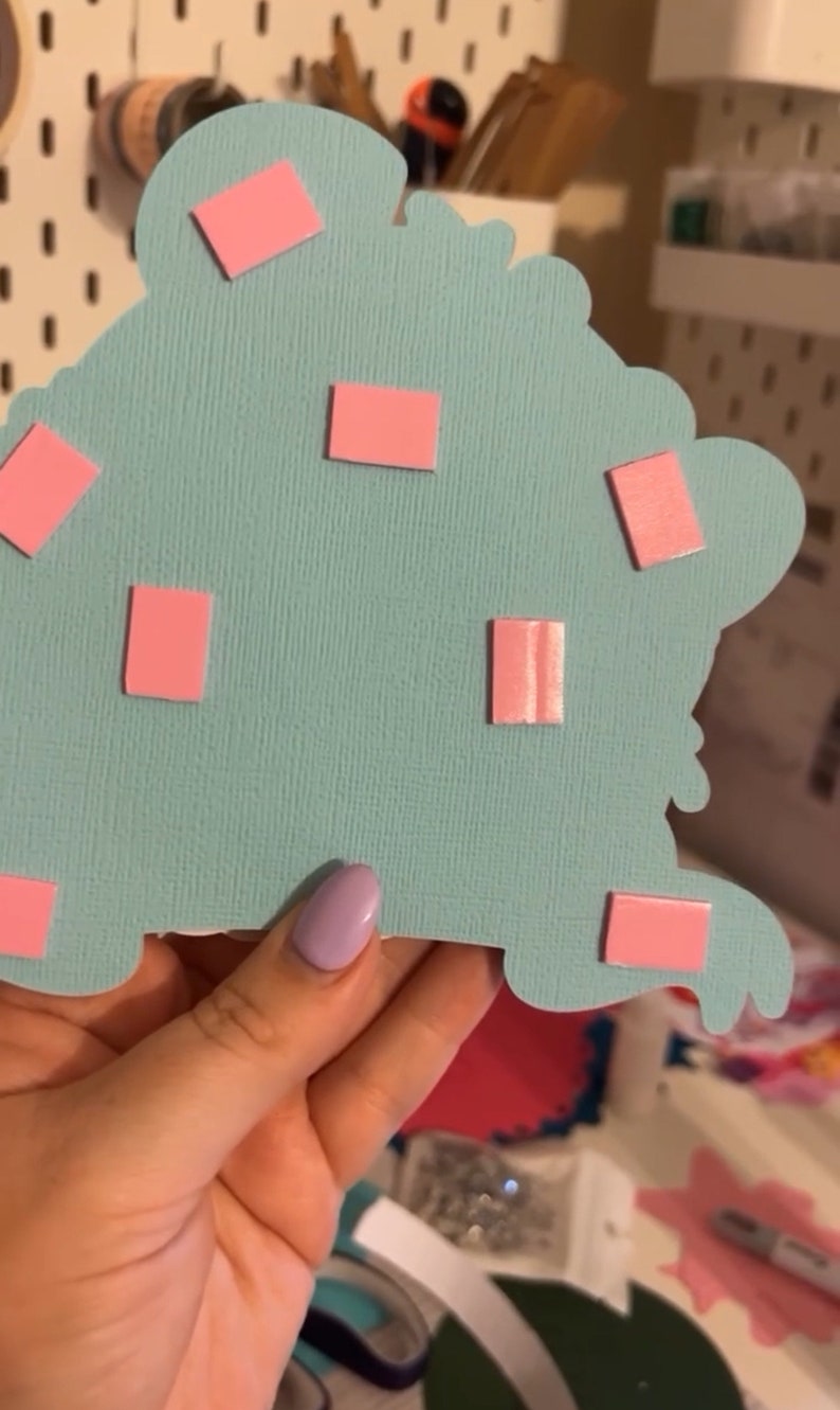 May include: A light blue paper craft with pink adhesive squares attached to the back. The craft is being held by a hand.