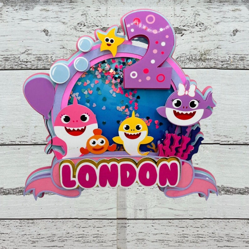 Peut inclure: D&eacute;coration de g&acirc;teau color&eacute;e avec des personnages de requins de dessins anim&eacute;s, une &eacute;toile de mer et le chiffre 2. Le design utilise des couleurs rose, violet et bleu, avec le nom "LONDON" affich&eacute;. Id&eacute;al pour un anniversaire.