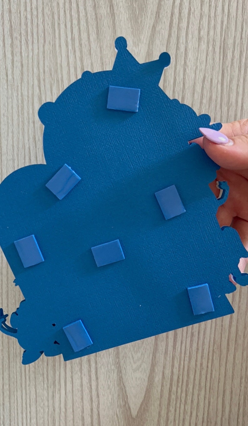 May include: Blue cardboard cutout of a castle with a crown on top. The back of the cutout has several adhesive squares attached to it.