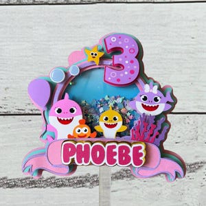Peut inclure: D&eacute;coration de g&acirc;teau multicouche avec un design rose et violet, le nom "PHOEBE" en lettres roses, des requins de dessin anim&eacute;, une &eacute;toile de mer et le chiffre "3". Le fond a un th&egrave;me oc&eacute;an bleu.