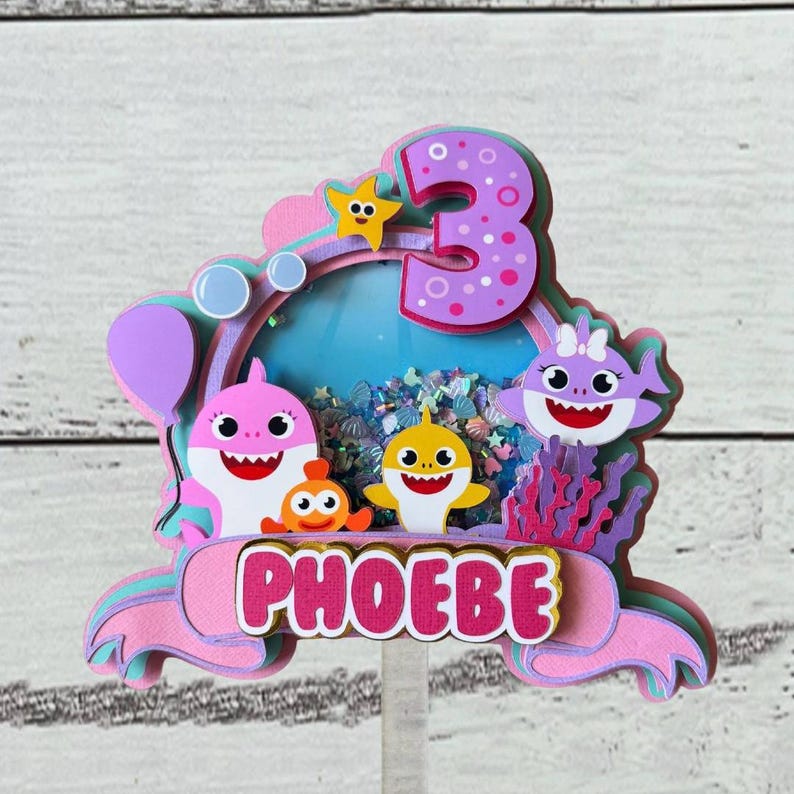 Peut inclure: D&eacute;coration de g&acirc;teau multicouche avec un design rose et violet, le nom "PHOEBE" en lettres roses, des requins de dessin anim&eacute;, une &eacute;toile de mer et le chiffre "3". Le fond a un th&egrave;me oc&eacute;an bleu.