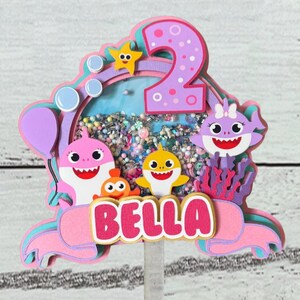 Peut inclure: Un cake topper multicouche rose, violet et bleu avec le nom "BELLA". Il pr&eacute;sente des personnages de requins de dessins anim&eacute;s, une &eacute;toile de mer et le chiffre "2". Le centre a une section transparente remplie de confettis color&eacute;s.