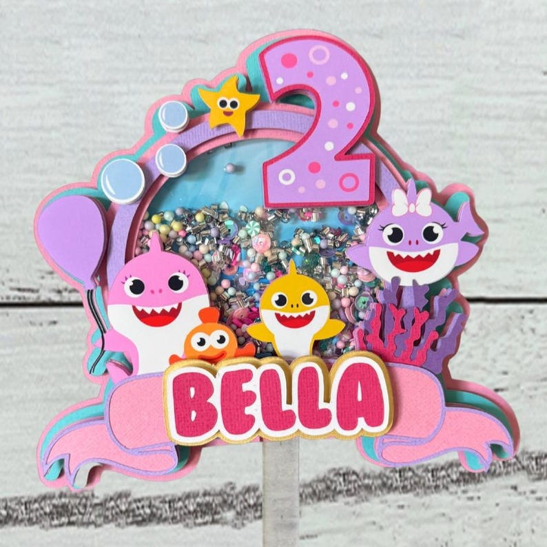Peut inclure: Un cake topper multicouche rose, violet et bleu avec le nom "BELLA". Il pr&eacute;sente des personnages de requins de dessins anim&eacute;s, une &eacute;toile de mer et le chiffre "2". Le centre a une section transparente remplie de confettis color&eacute;s.