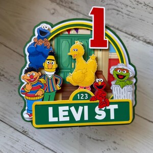 May include: Sesame Street themed cake topper with Big Bird, Elmo, Cookie Monster, Bert and Ernie, and Oscar the Grouch. The topper features a green street sign with the text "LEVI ST" and the number "123".