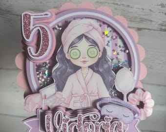 Op de afbeelding: Een gelaagde papieren taarttopper met een glitterend cijfer 5 en de naam Victoria in roze schrift. Het midden toont een illustratie van een persoon in een badjas met komkommerschijfjes op de ogen, omringd door kralen en pailletten.
