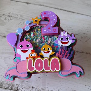 Peut inclure: D&eacute;coration de g&acirc;teau multicouche avec des personnages de requins de dessins anim&eacute;s, une &eacute;toile de mer et le chiffre 2. Le nom "LOLA" est affich&eacute; en lettres roses. La d&eacute;coration a un b&acirc;ton en plastique transparent.