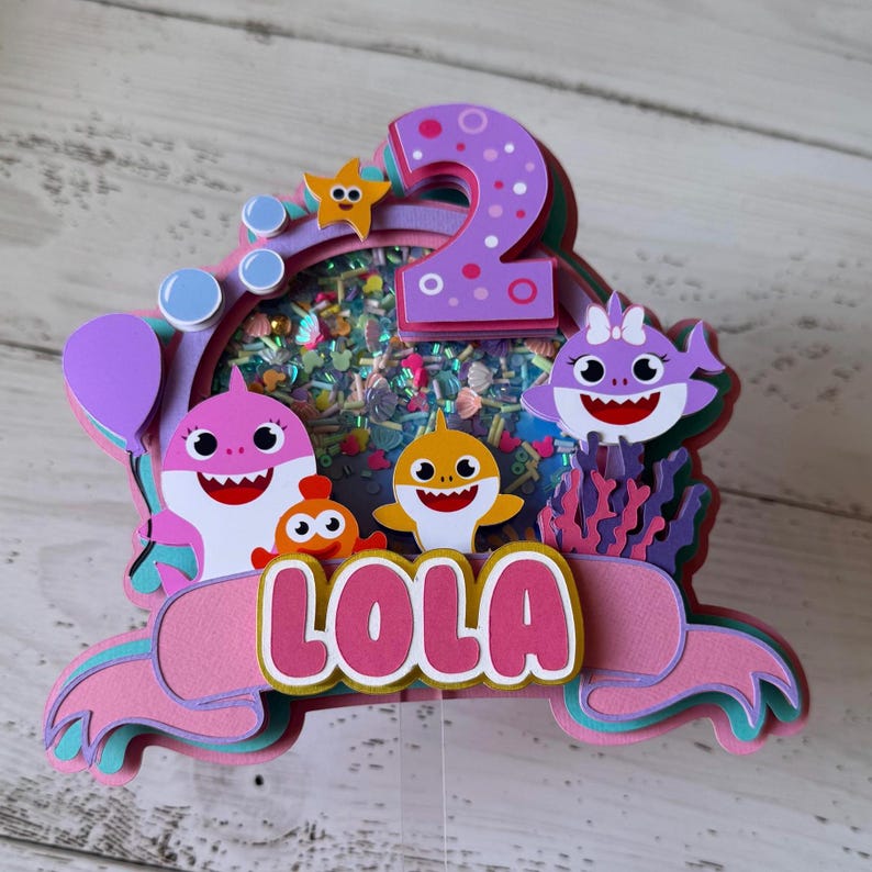 Peut inclure: D&eacute;coration de g&acirc;teau multicouche avec des personnages de requins de dessins anim&eacute;s, une &eacute;toile de mer et le chiffre 2. Le nom "LOLA" est affich&eacute; en lettres roses. La d&eacute;coration a un b&acirc;ton en plastique transparent.