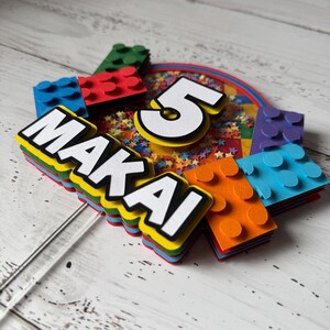 Op de afbeelding: Een kleurrijke taarttopper met een Lego-thema. De topper heeft een nummer 5, de naam Makai en kleurrijke Lego-stenen.