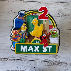 May include: A Sesame Street themed cake topper with a green, yellow, and red color scheme. The topper features the number 2, Big Bird, Elmo, Cookie Monster, and a trash can. The topper also includes the text "Max St" and "123".