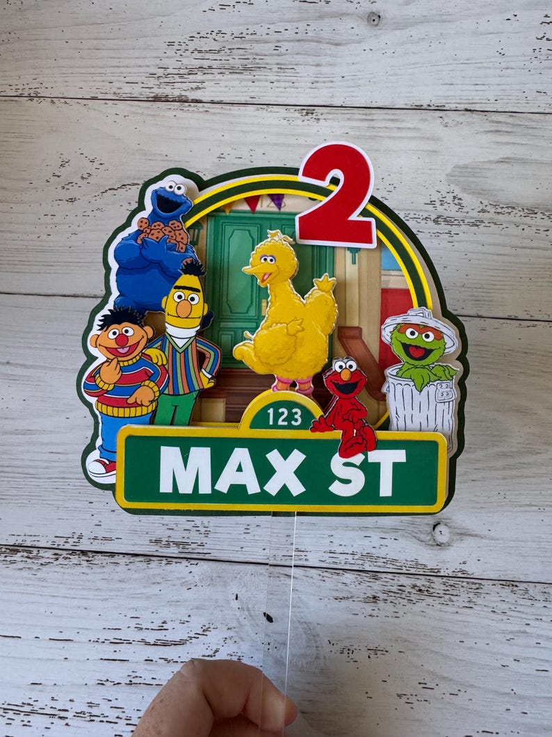 May include: A Sesame Street themed cake topper with a green, yellow, and red color scheme. The topper features the number 2, Big Bird, Elmo, Cookie Monster, and a trash can. The topper also includes the text "Max St" and "123".