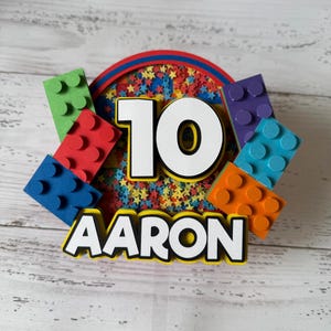 Op de afbeelding: Een kleurrijke verjaardagstaarttopper met het nummer 10 en de naam Aaron. De topper is versierd met kleurrijk confetti en Lego-blokken.