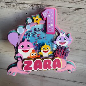 Peut inclure: D&eacute;coration de g&acirc;teau multicouche avec des personnages de requins de dessins anim&eacute;s, une &eacute;toile de mer et le chiffre 1. Le nom "ZARA" est affich&eacute;. La d&eacute;coration est principalement rose, violet et bleu, avec un &eacute;l&eacute;ment vibrant.