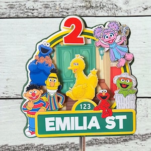 May include: A Sesame Street themed cake topper with Big Bird, Elmo, Cookie Monster, Bert, Ernie, and a pink character holding a wand. The topper features a green street sign that says "EMILIA ST" and the number "123". The number "2" is also featured on the topper.
