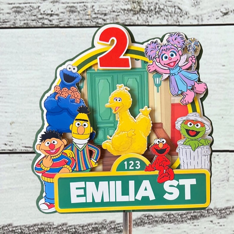 May include: A Sesame Street themed cake topper with Big Bird, Elmo, Cookie Monster, Bert, Ernie, and a pink character holding a wand. The topper features a green street sign that says "EMILIA ST" and the number "123". The number "2" is also featured on the topper.